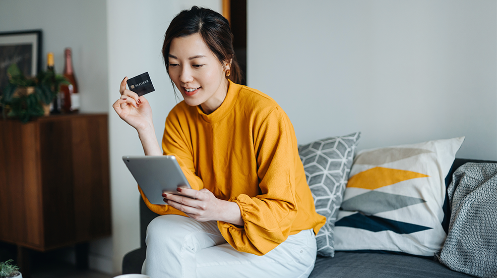 Beautiful smiling young Asian woman with credit card managing personal banking and finance with online banking mobile app on digital tablet at cozy home